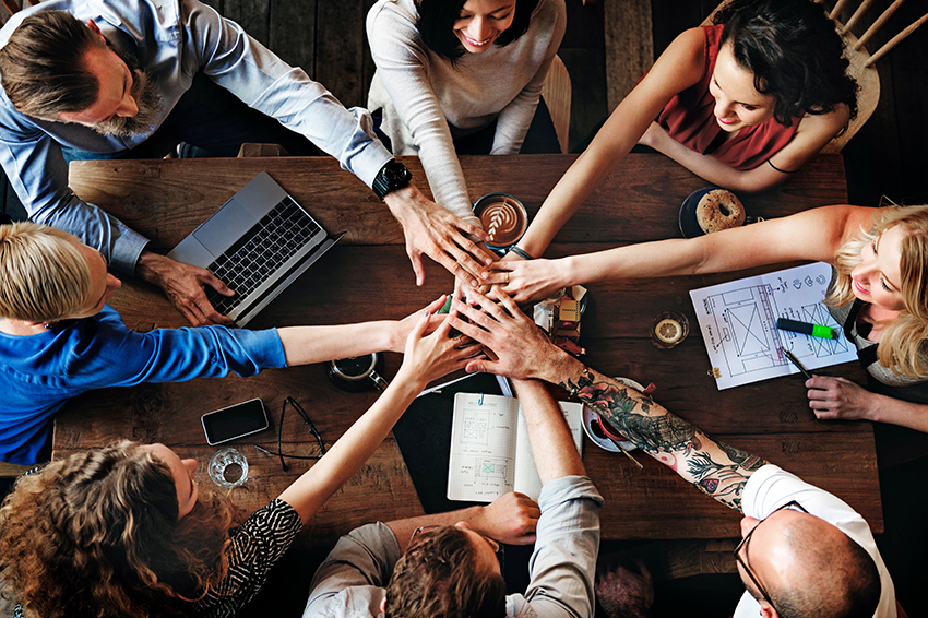 Das Titelbild des Blogs über das Employer Branding Teil 2 zeigt ein Team, was an einem im Kreis um einen Tisch sitzt und die Hände in der Mitte zusammenhält.